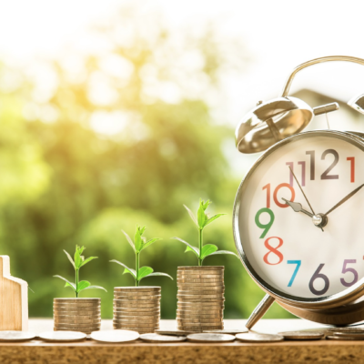 A wooden model of a house, an increasing number of coins with small plants, and an alarm clock.