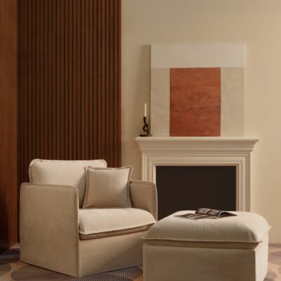 Anabei armchair and matching ottoman in Mist, a soft beige washable fabric, beside a white fireplace